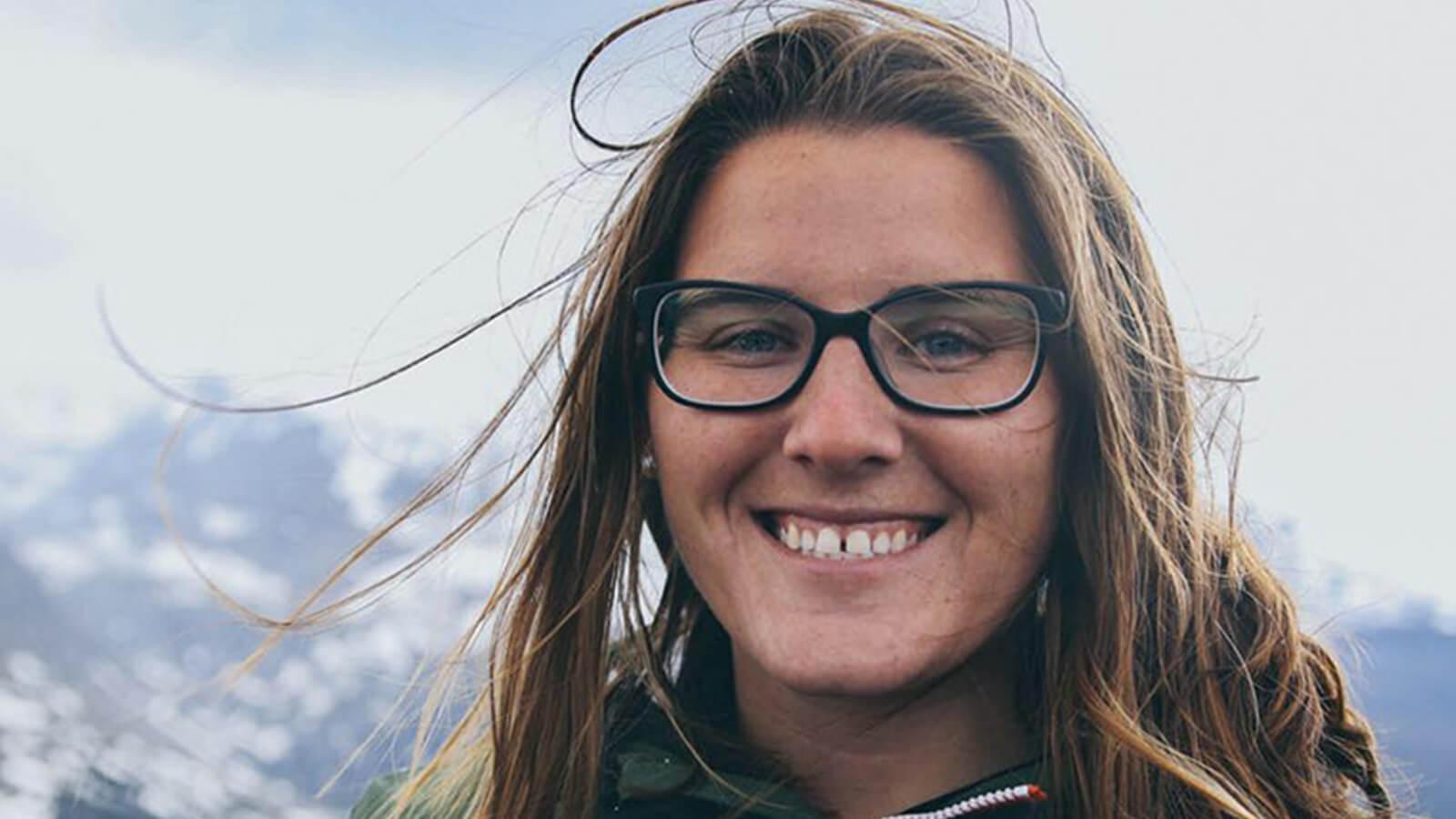 Full Sail grad SaJorden Miller pictured from the shoulders up, in the background is a snowcapped mountain range. She is smiling, wearing a green zip-up jacket with orange accents and black framed glasses.