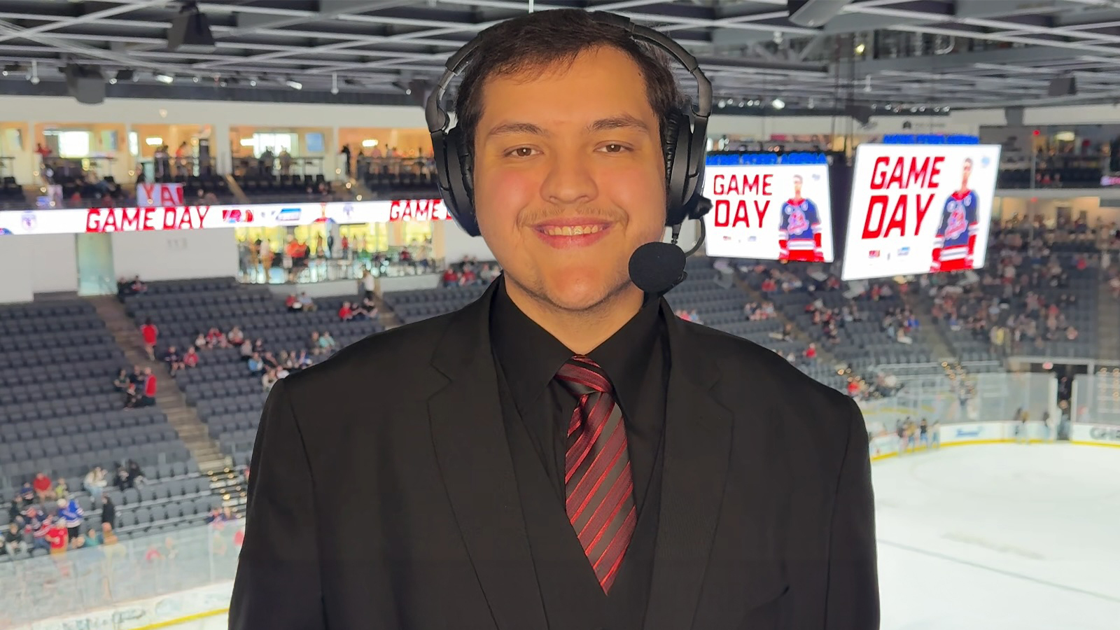 A smiling sports broadcaster in a suit and headset stands in an arena booth above an ice hockey rink with "Game Day" on screens behind.