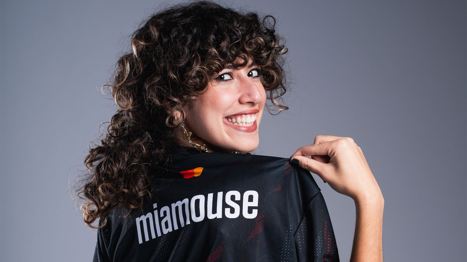 A smiling woman with curly hair holds up a jersey that reads “miamouse” on the back, showing it proudly to the camera.