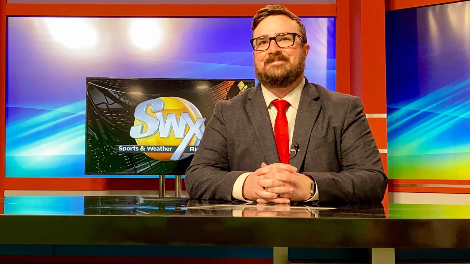 Richard Tieman wears a suit and sits at a TV broadcast desk. A screen behind him says SWX Sports and Weather.