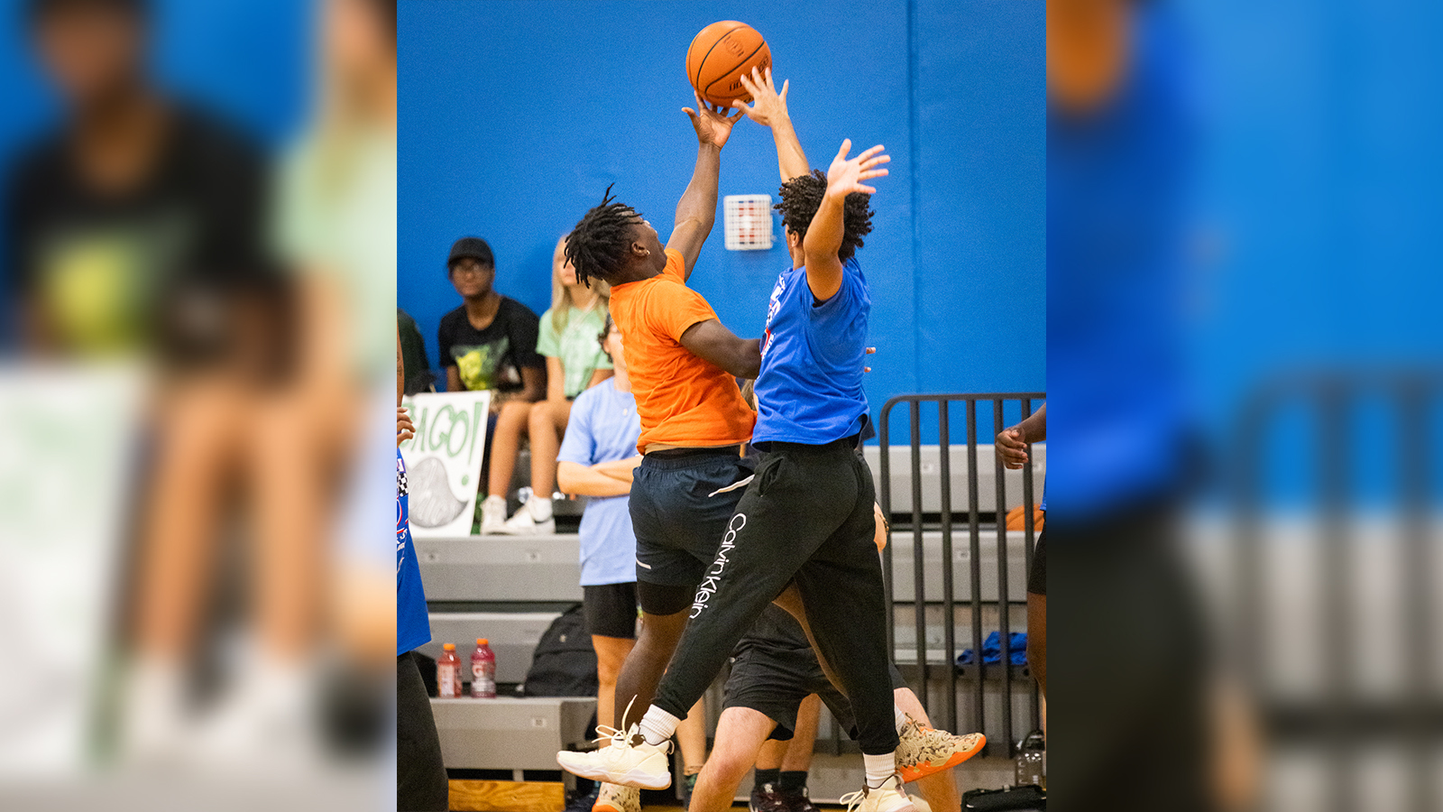 Two basketball players jump up simultaneously to reach the basketball on a court. They are both wearing athletic clothing.