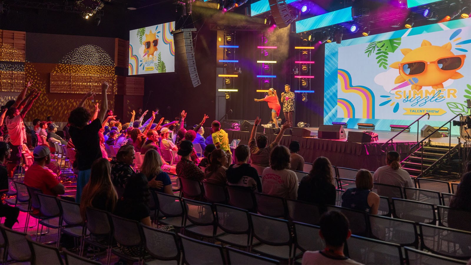 Several students raise their hands attentively at the Summer Sizzle as two staff members call on participants. The large room is colorfully lit up and looks very lively.