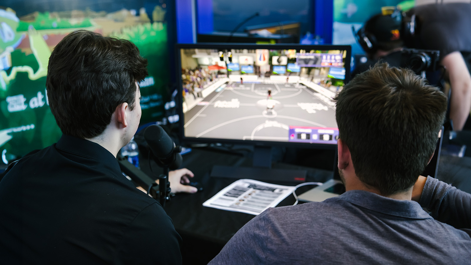 Two people have their backs to the camera while watching ‘NBA 2K’ gameplay on a small monitor on a table in front of them.