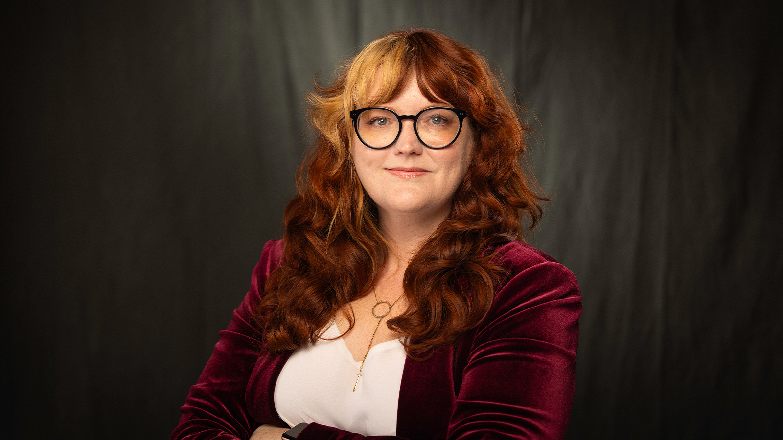 Susan Kost smiles at the camera. She has long red hair and is wearing a white blouse, a red velvet jacket, and black glasses.