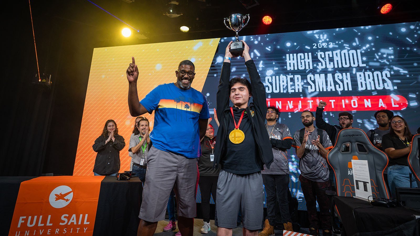 Two people are standing side by side on a stage while one holds a large silver trophy over their head.