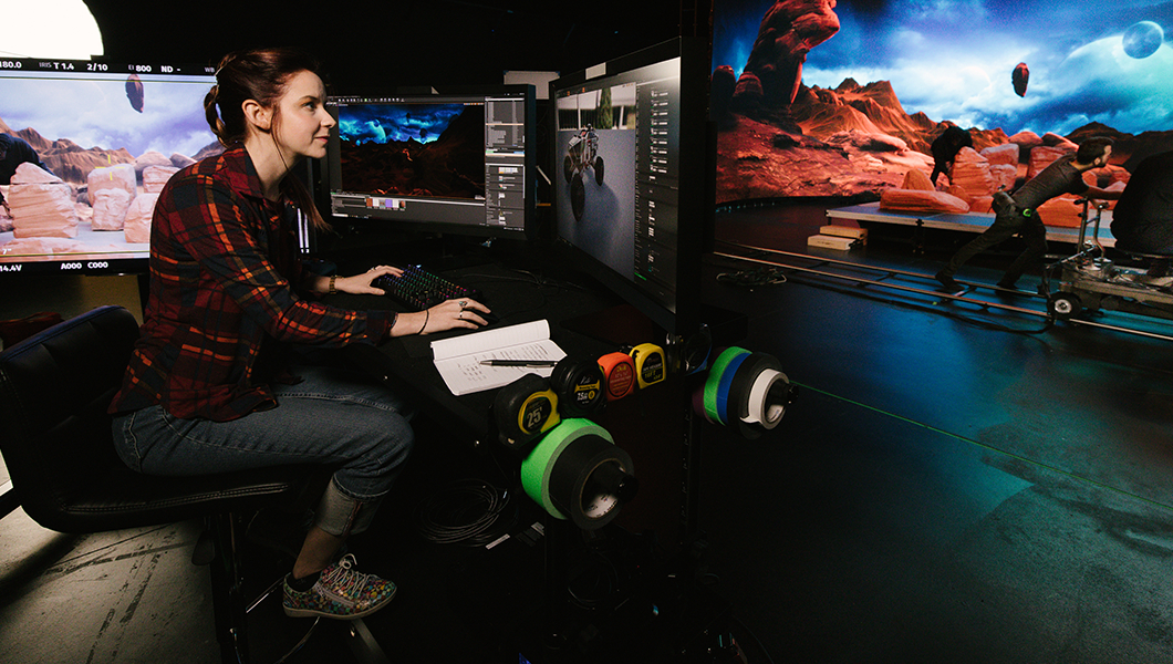 A woman is sitting at a desk in front of several monitors. She is in a virtual production studio that has been designed to look similar to Mars.