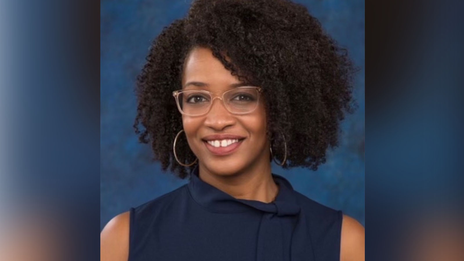 Tiana, an African American woman, sits smiling at the camera. She is wearing a sleeveless navy blue blouse, silver hoop earrings, and glasses with translucent beige frames.
