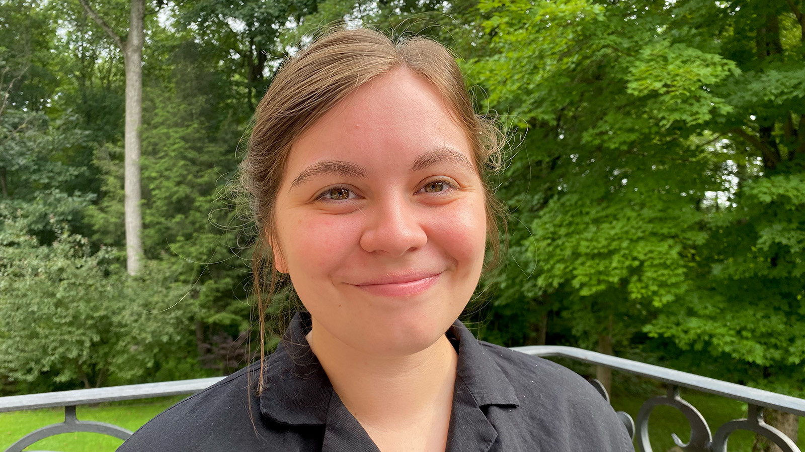 Emmi Litsenberger sits on a porch in front of a grassy lawn and trees. She is smiling and wearing a gray button-up shirt.