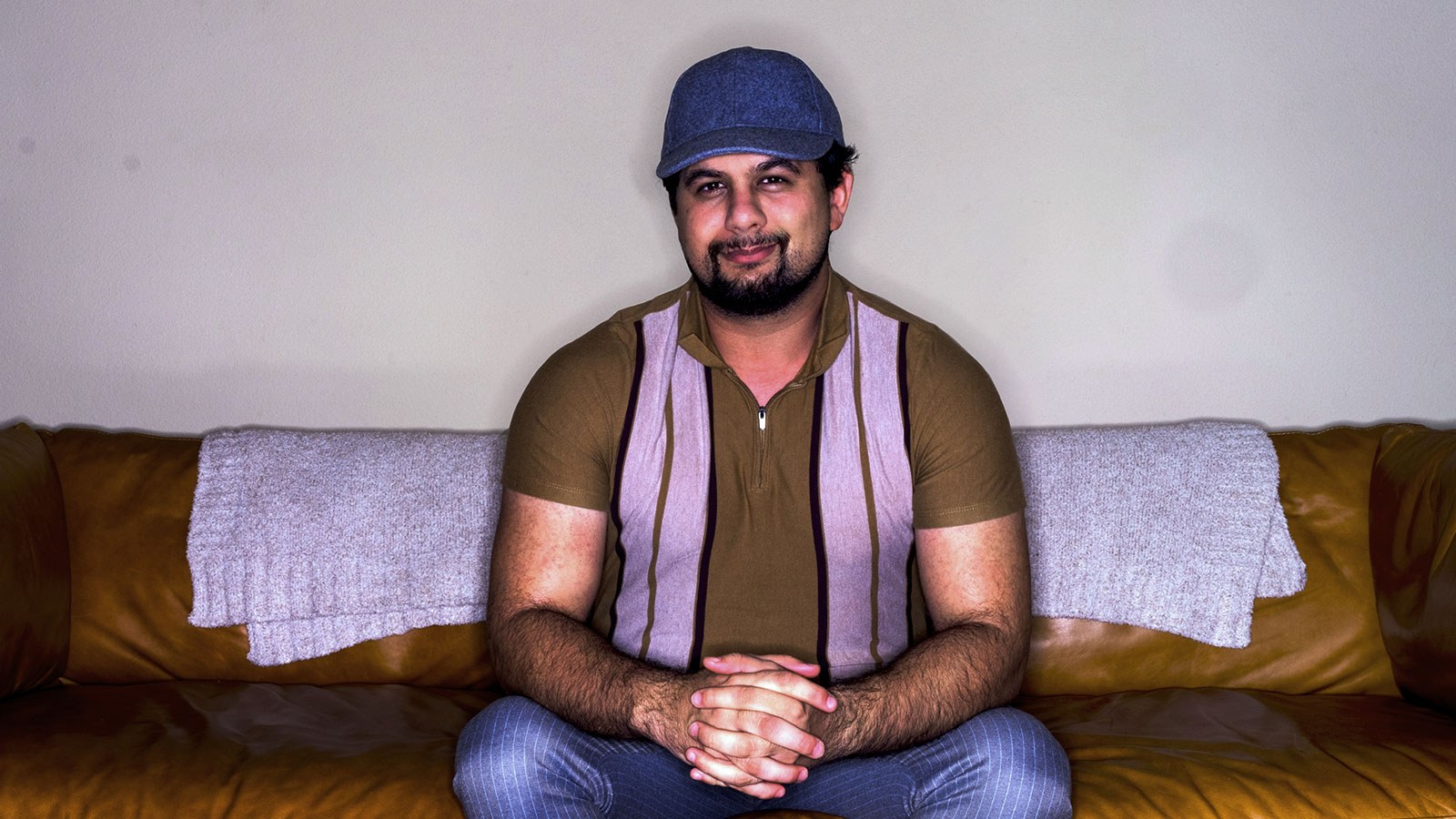 Tripp Topping sits on a leather couch. He is wearing a brown half-zip shirt, blue pinstriped pants, and a blue baseball cap.