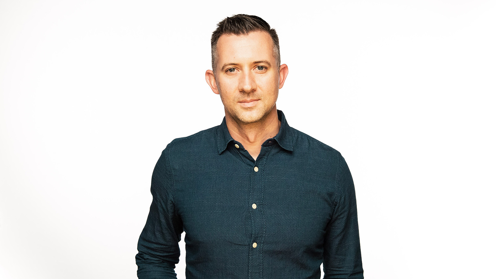 Dean Sell stands in front of a white background. He is wearing a dark button down shirt.