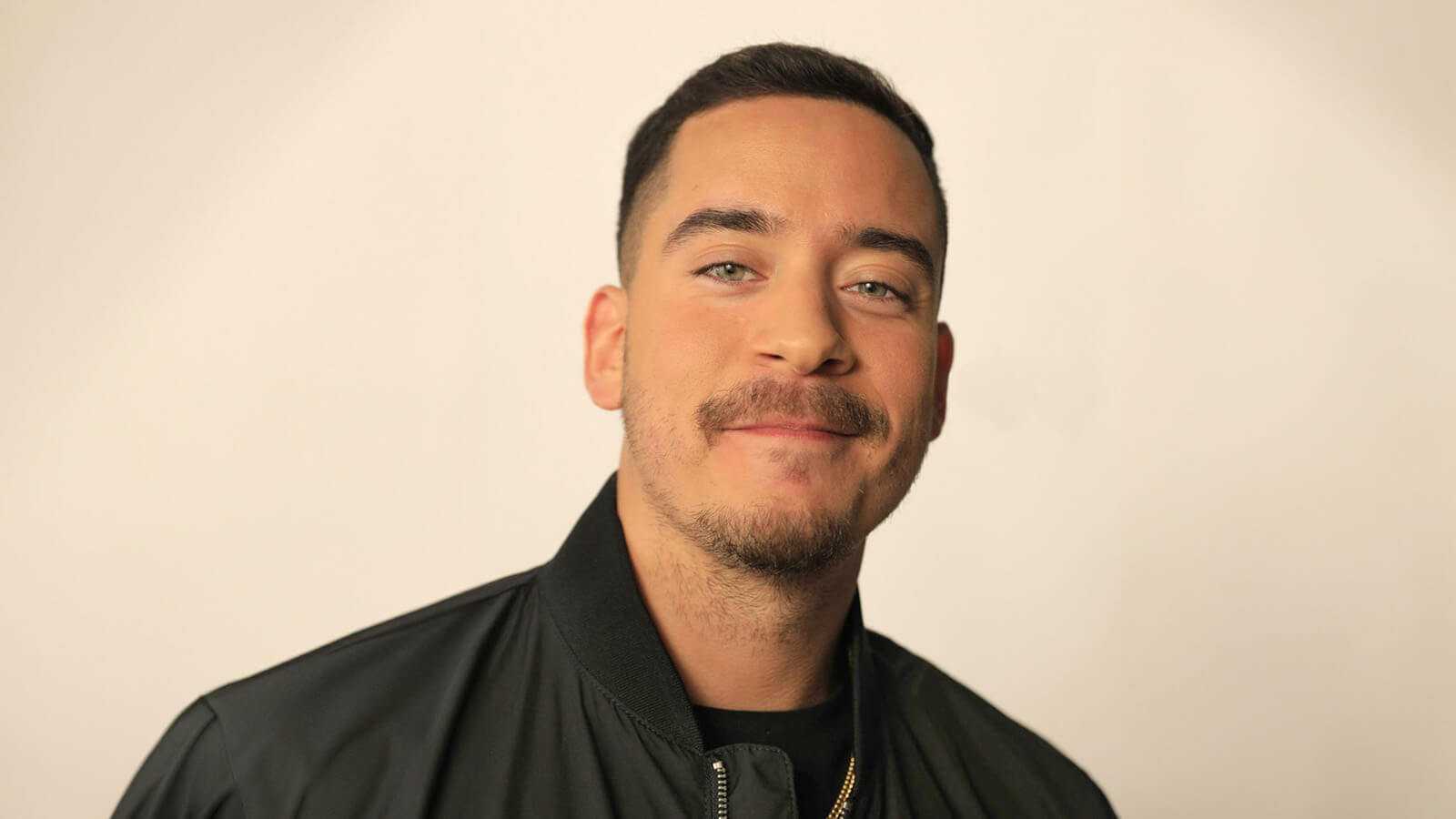 Eric Medina smiles at the camera. He is wearing a black zip-up jacket, black t-shirt, and gold chain necklace.