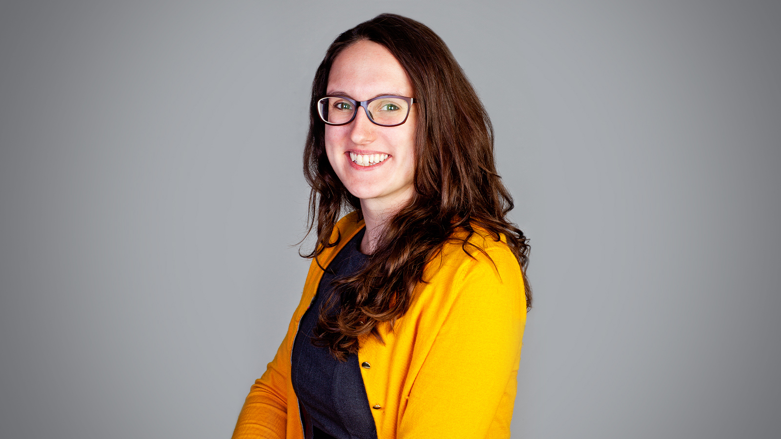 Tasha Rosenbaum smiles at the camera. She has brown hair and is wearing glasses, a yellow cardigan, and a blue sweater.