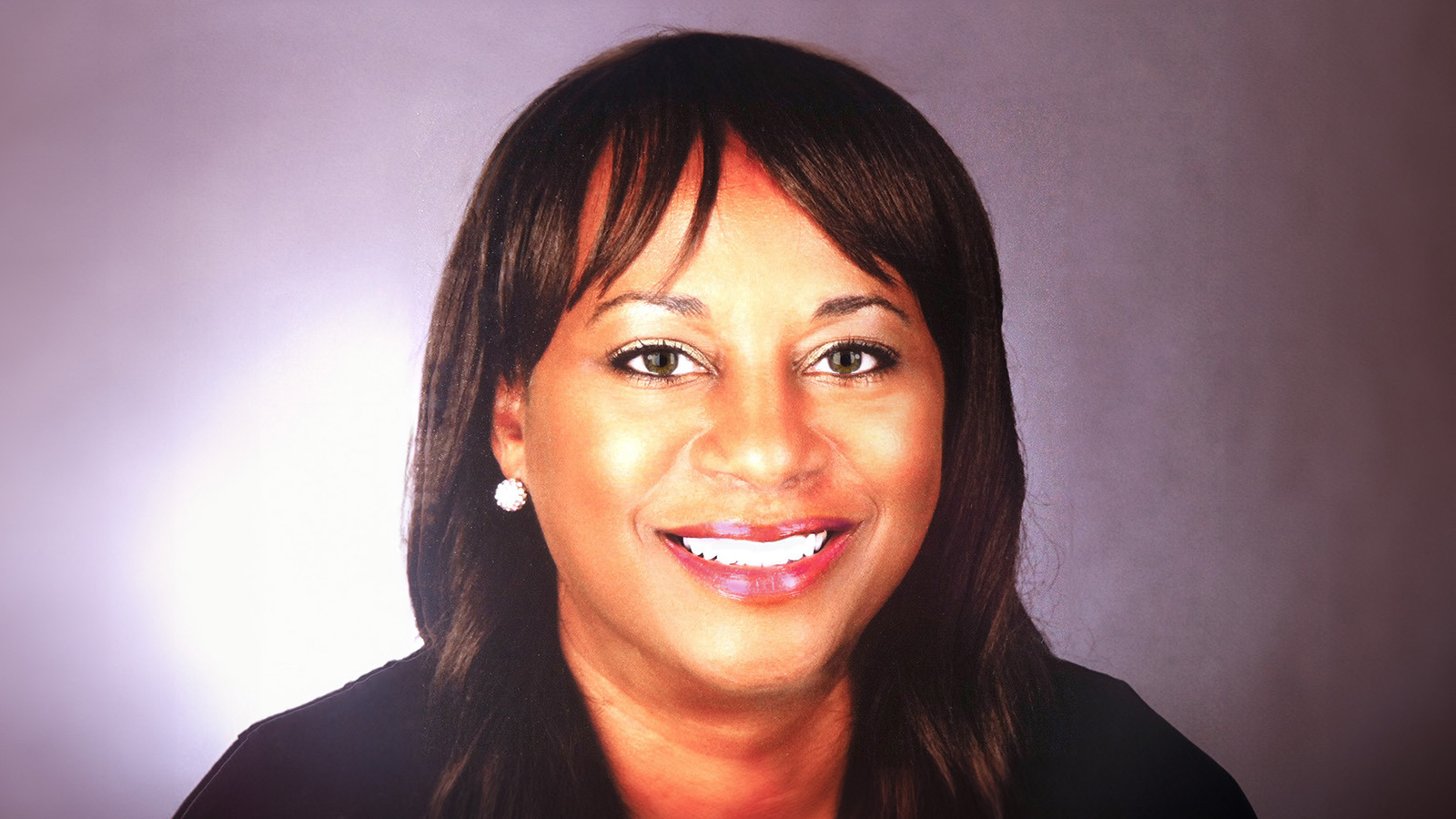 Lorraine smiles at the camera. She has dark brown hair and hazel eyes and is wearing a blue blazer and silver earrings.