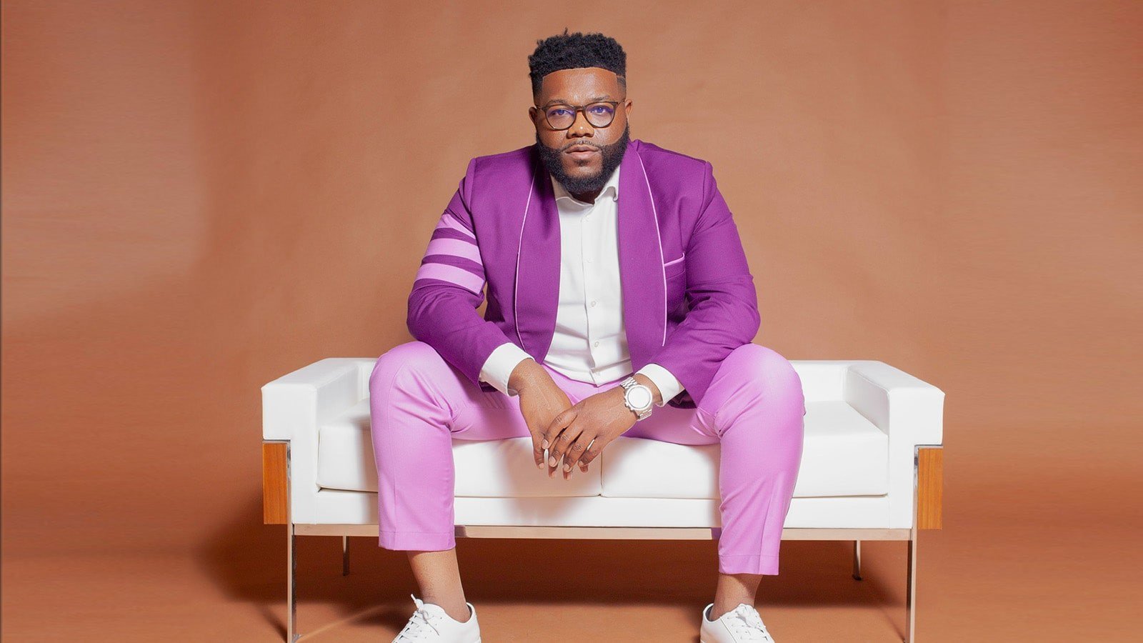 A man with black hair and a beard wearing glasses, a purple suit, and a white button down shirt while seated on a white couch against a brown background.