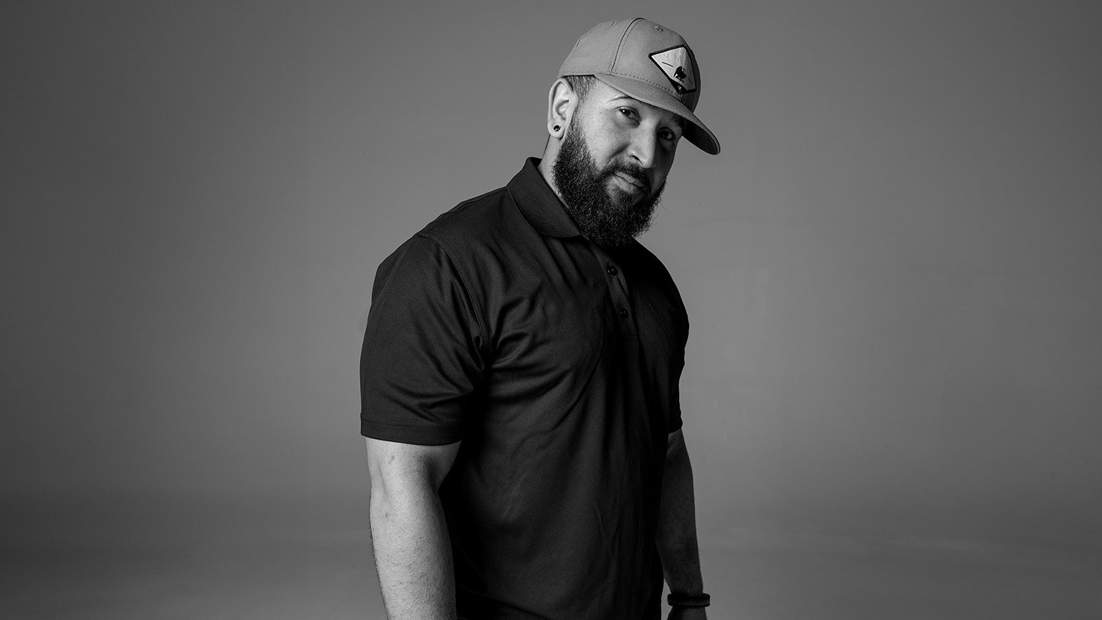 Kevin Hall stands and looks at the camera. He has a beard and is wearing a baseball cap, a collared short-sleeve shirt, and jeans.