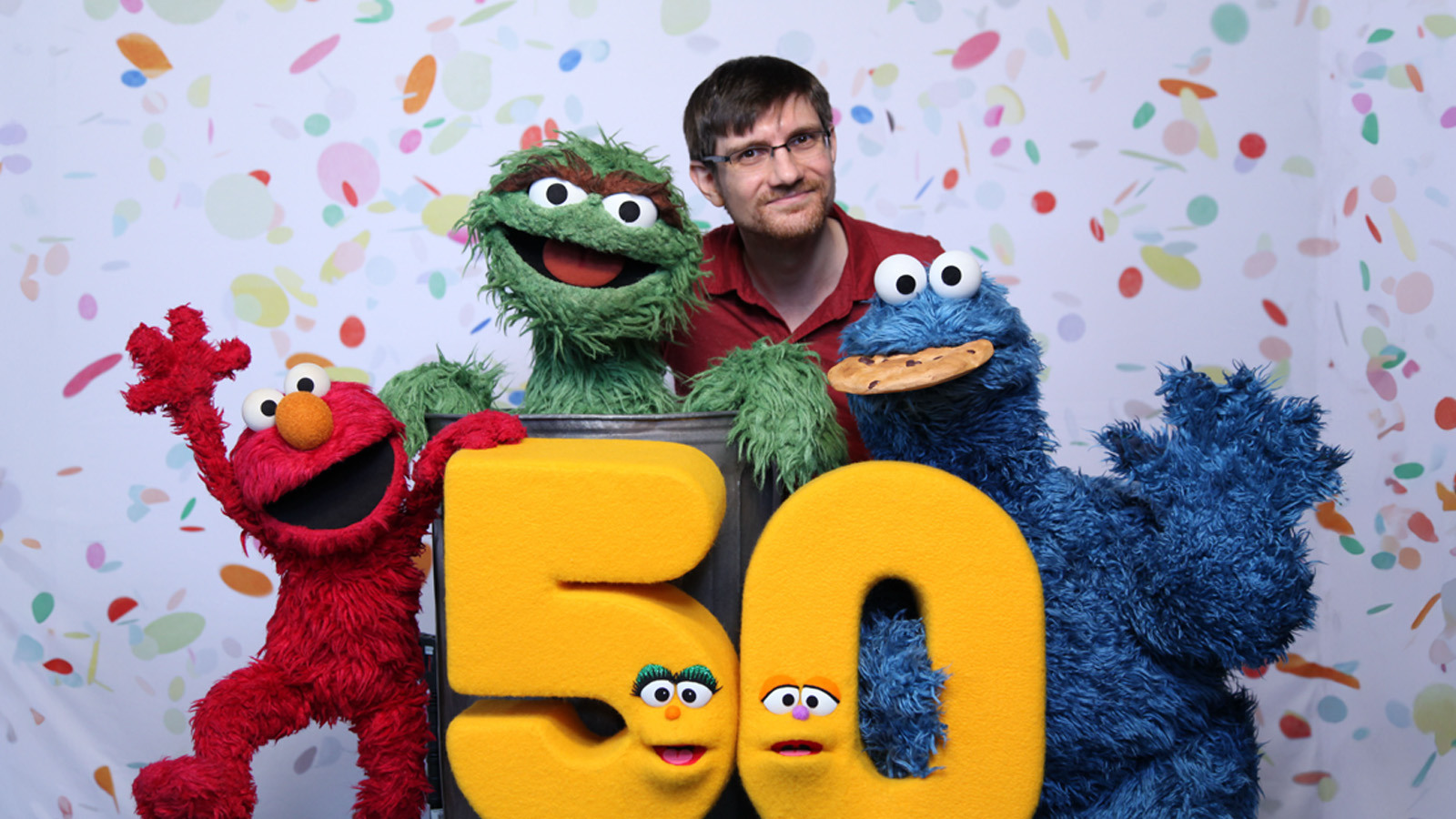 Chris Sassano stands with Oscar the Grouch, Cookie Monster, Elmo, and a puppet shaped like the number 50 with confetti behind them.
