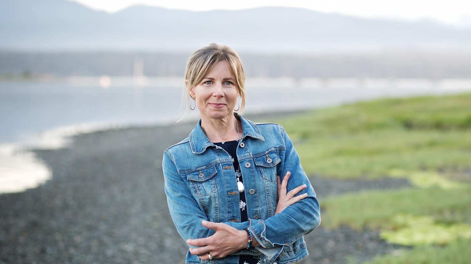 A blonde woman wearing a denim jacket over a navy top and black skirt stands with her arms crossed. She is outdoors in front of a river with mountains far in the background.