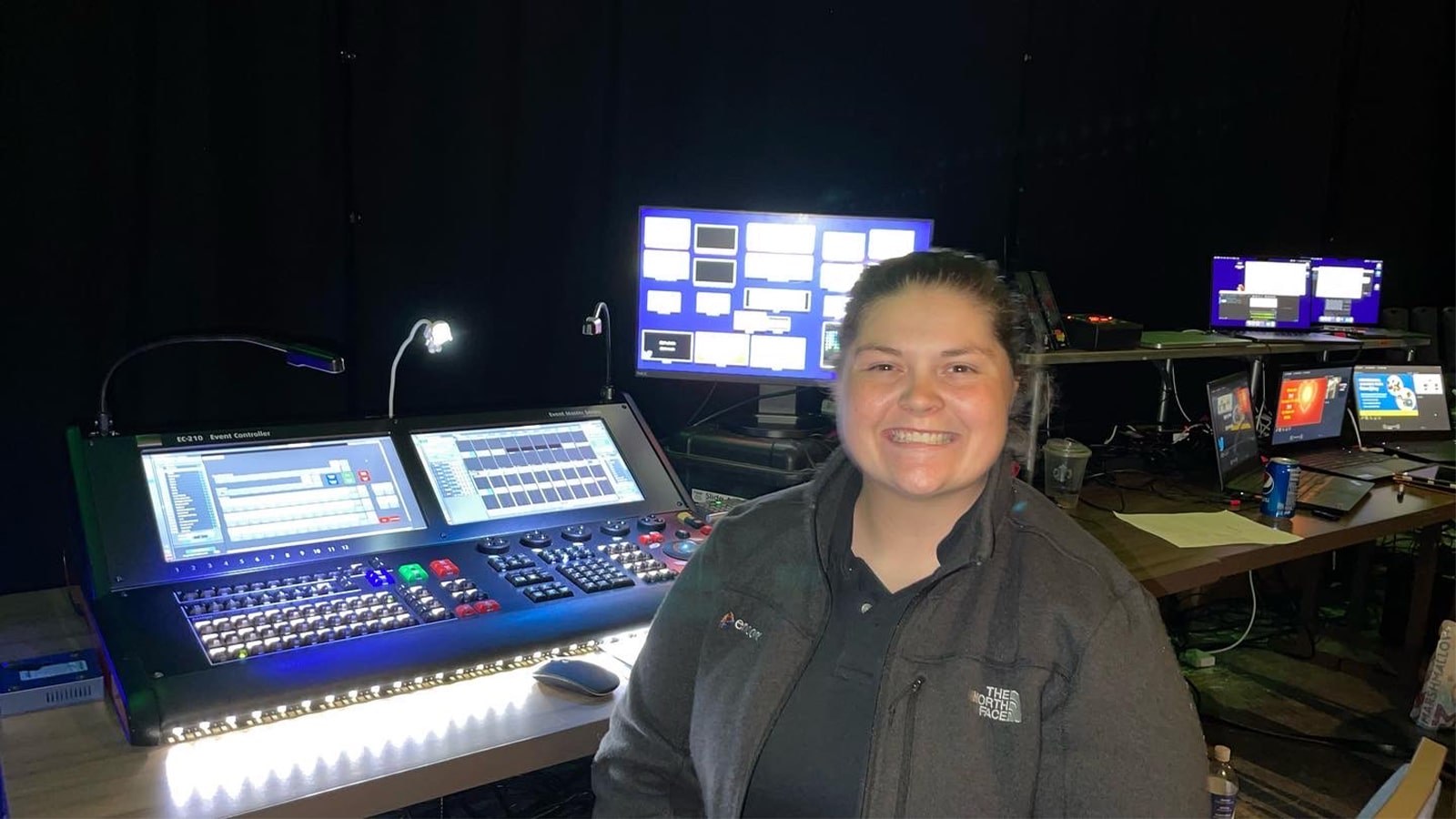 Jean Fuemmeler sits in front of live event video production controllers. She is wearing a gray jacket and smiling.