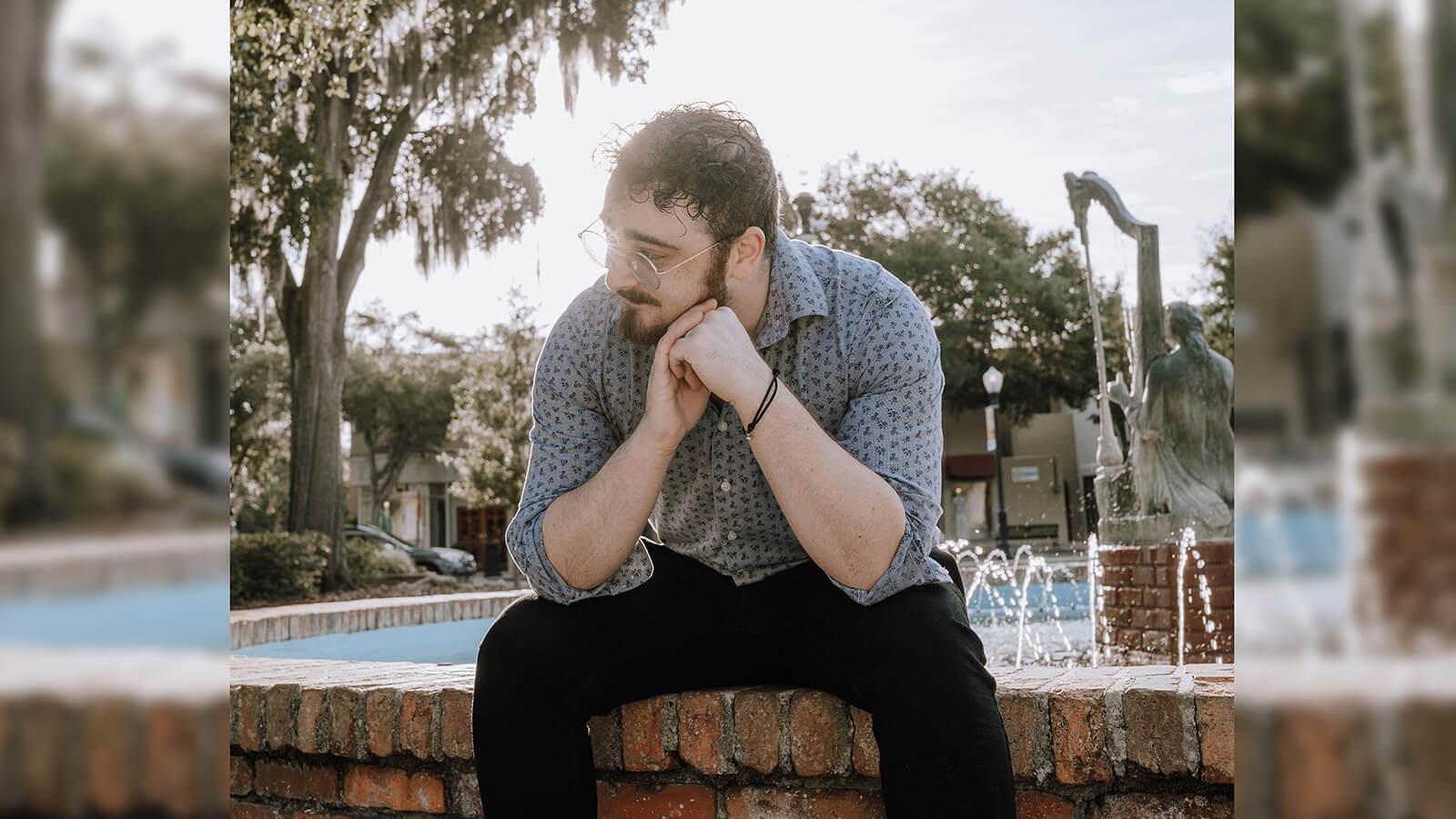 A man with short, dark, curly hair in a blue button down shirt and wireframe glasses looking to the side while sitting in front of a fountain.