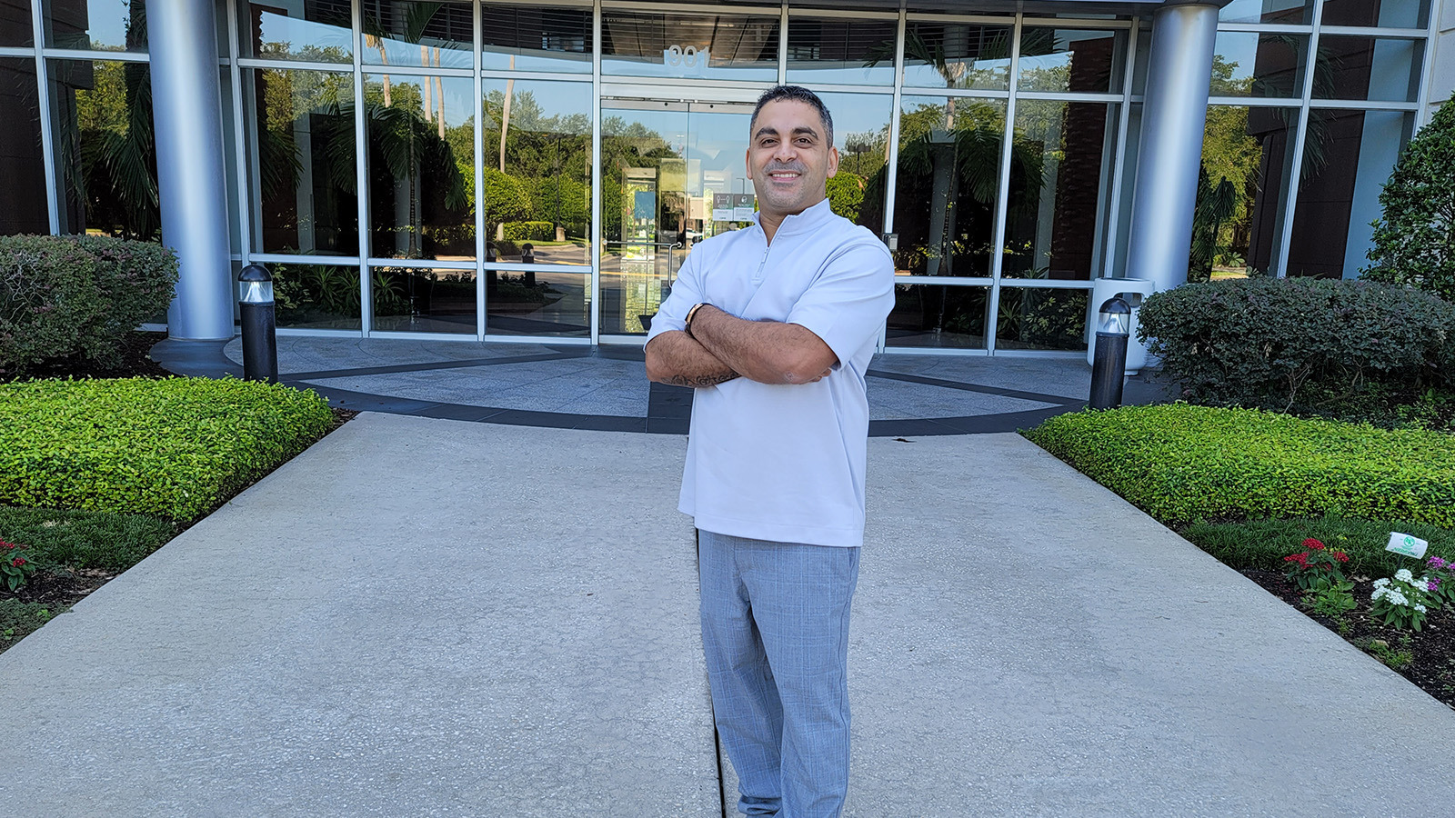 Brian Alvarado stands smiling with his arms crossed in front of the Deloitte building. He wears gray slacks and a blue shirt.