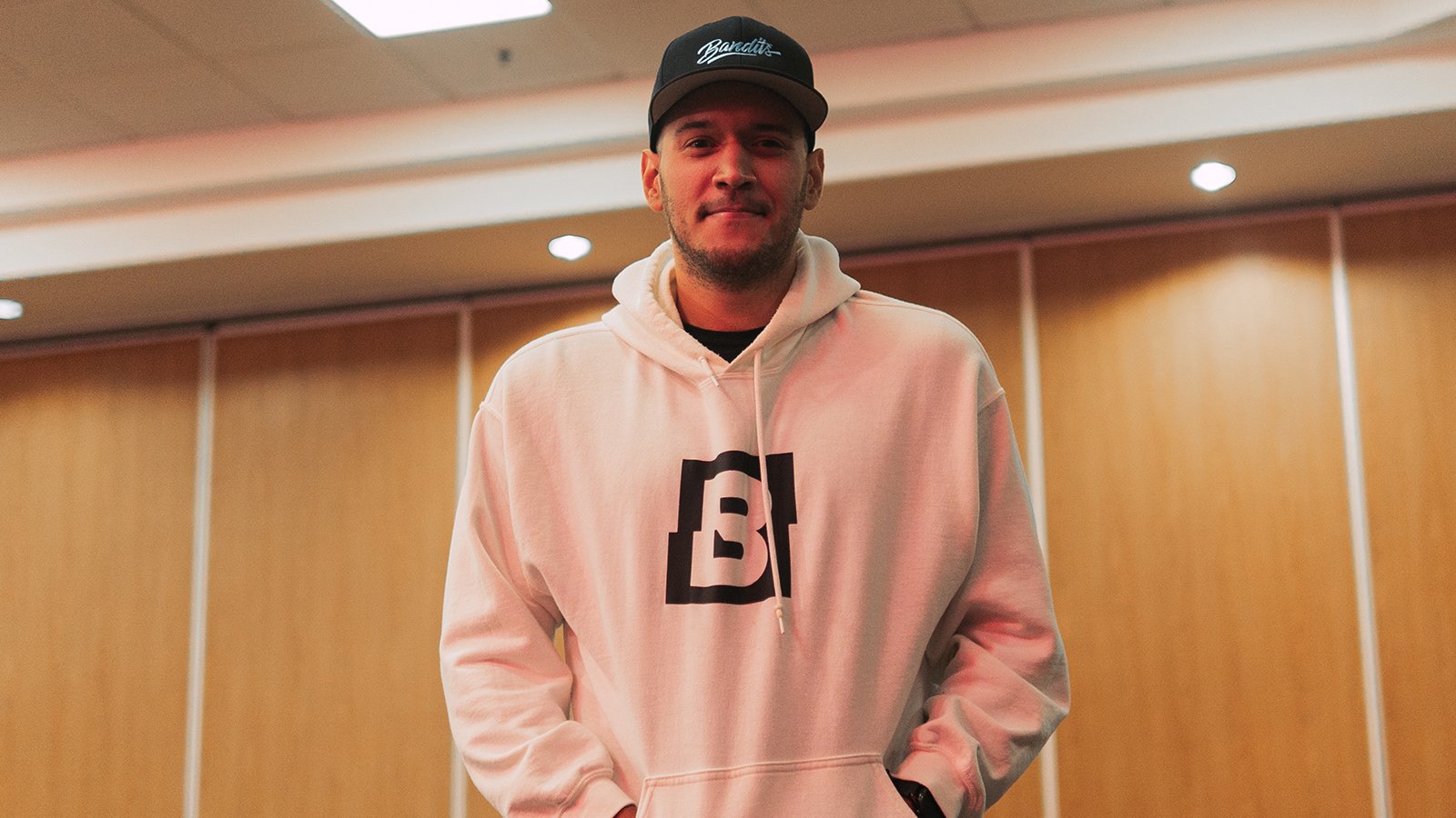 A man standing against a wood paneled wall in a white hooded sweatshirt featuring the Bandits logo and a black Bandits Gaming snapback cap.