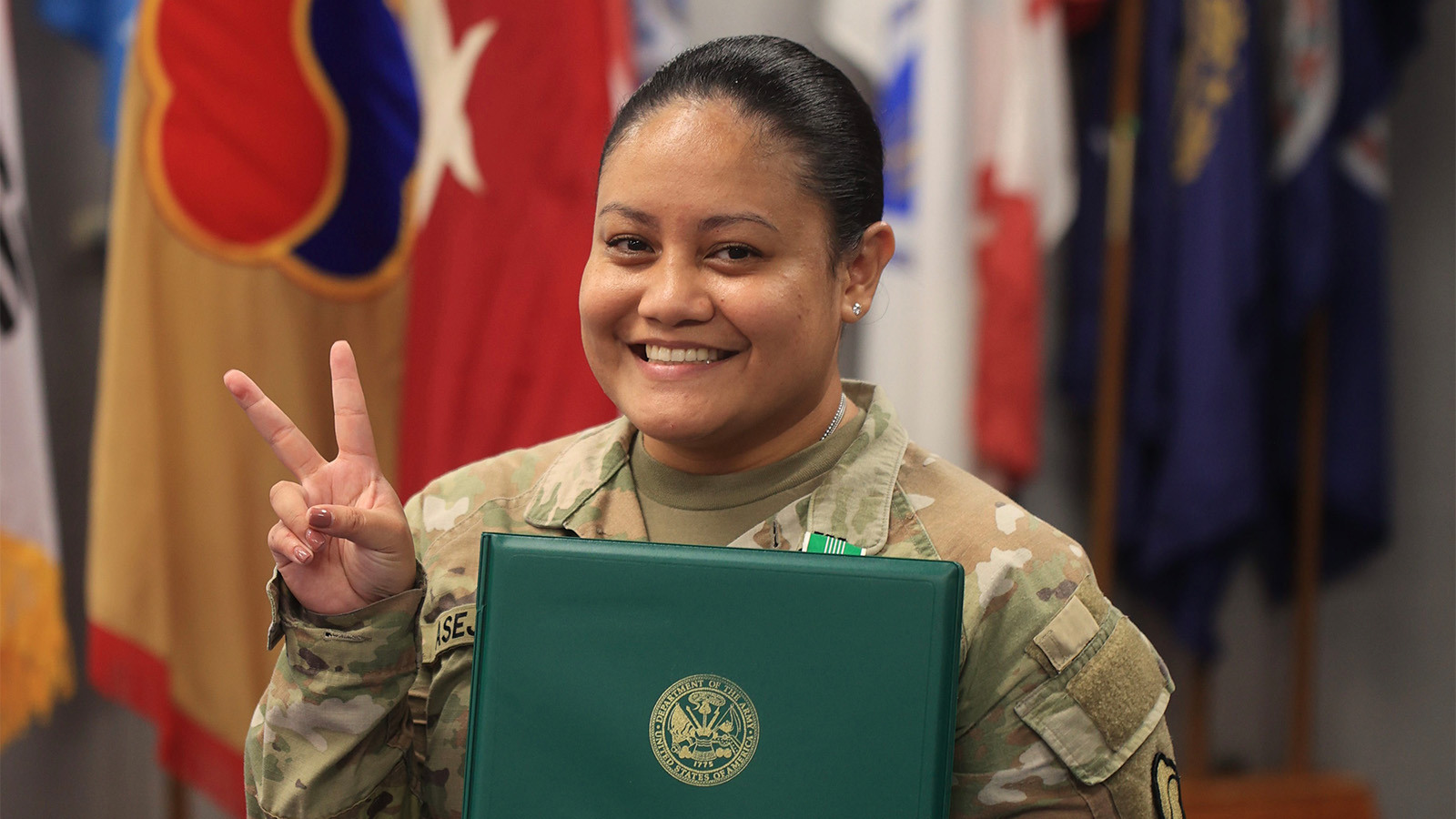 Anjelica Asejo smiles at the camera holding a certificate. She is wearing her Army uniform with a row of flags behind her as she holds her fingers in a peace sign.