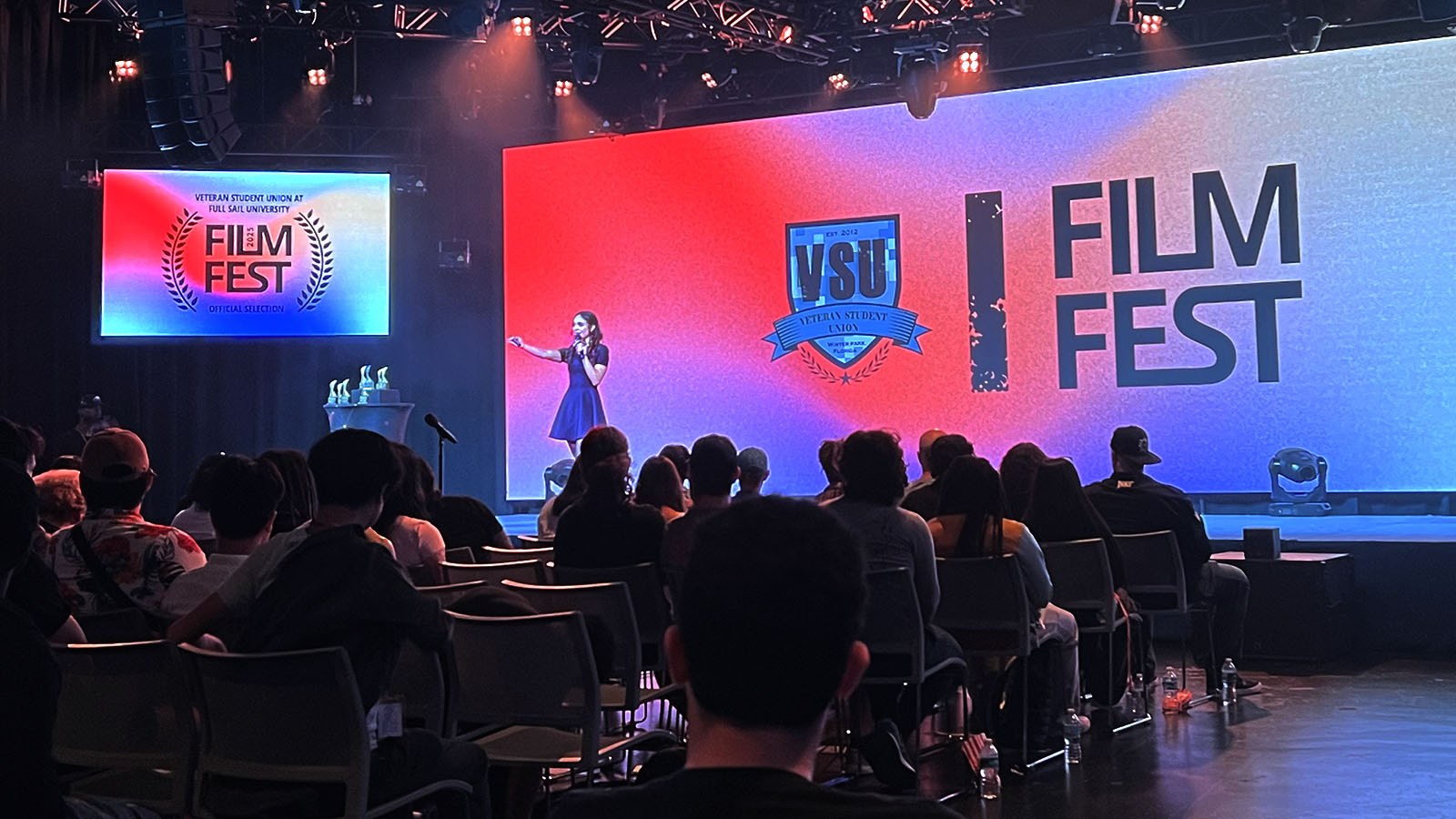 An audience faces a stage with a speaker holding a microphone. A large screen displays “Film Fest” and the VSU logo on a colorful background.