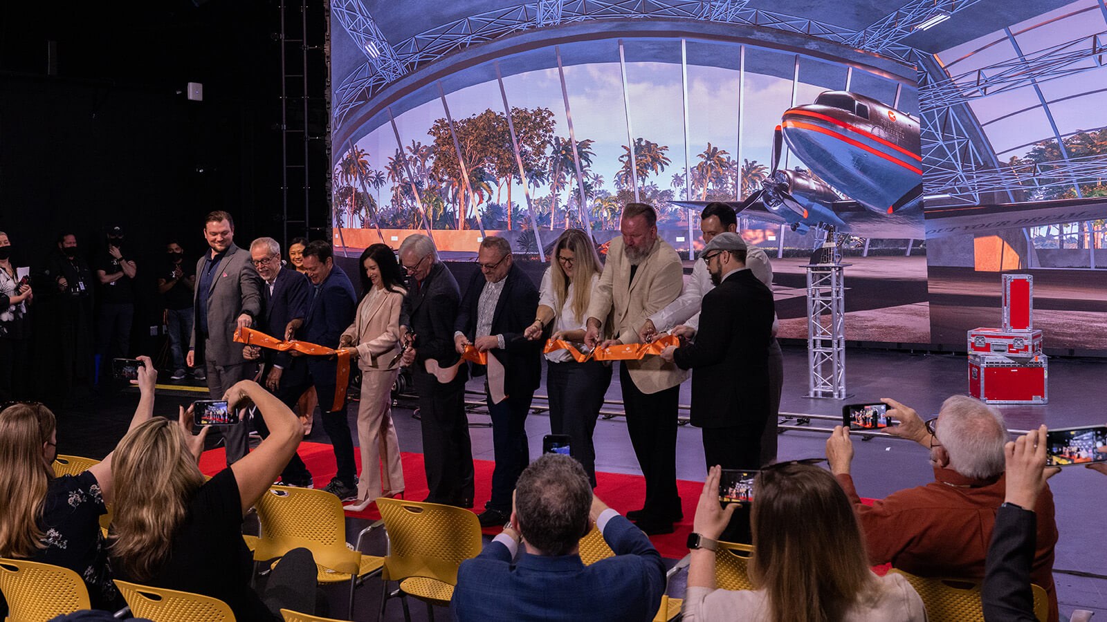 A group of eight men and two women stand on a red carpet in front of a large screen displaying an airplane and palm trees. They are each holding a pair of scissors and cutting a long orange ribbon. The seven audience members in front of them are photographing them with cell phones.