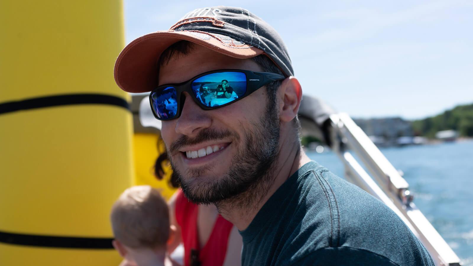 A man wearing a blue t-shirt, a blue and red baseball cap, and sunglasses with polarized lenses sits on a boat on a sunny day. He is smiling at the camera and there is a brunette woman and a blonde child sitting out of focus behind him.