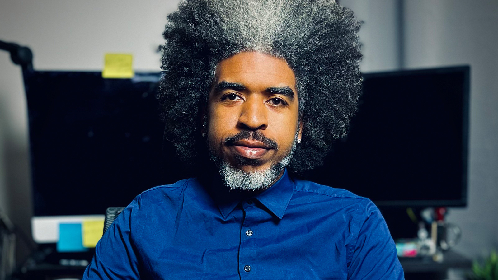 Bill Hudson sits in an office chair in front of a desk with two computer monitors. He has black and gray hair and wears a blue button-up shirt.