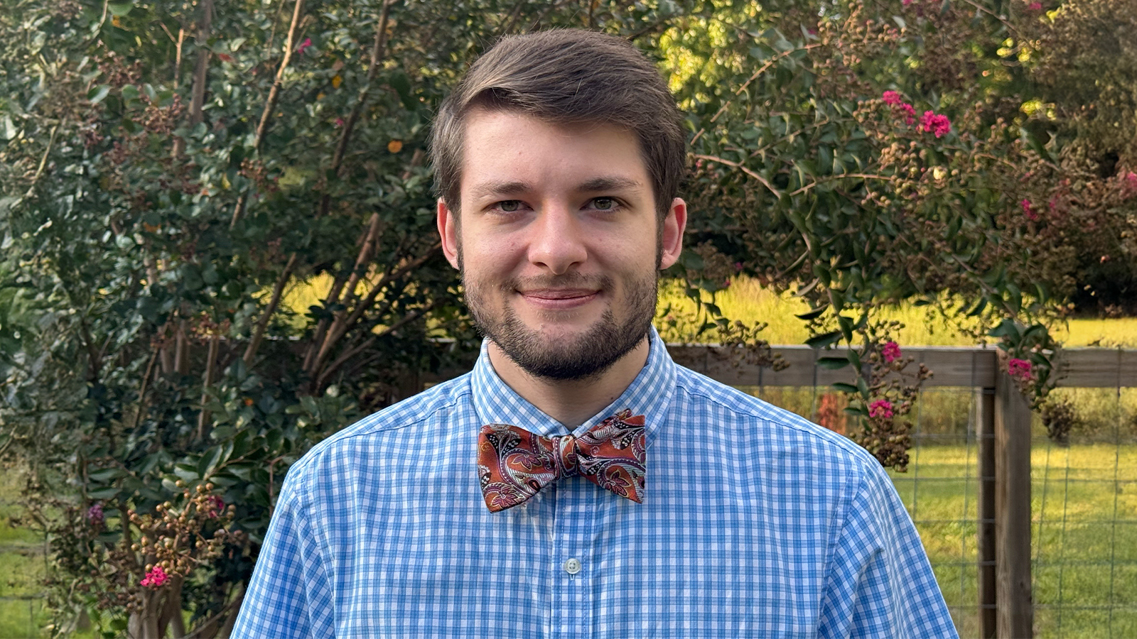 Noah White smiles at the camera in front of a row of green bushes with pink flowers. He is wearing a blue shirt and red bowtie.