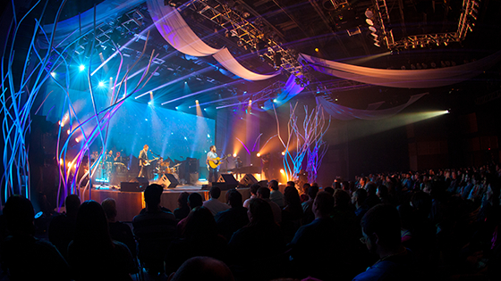 A band plays music in a large, dark auditorium with several bright blue and purple lights pointing at the stage.