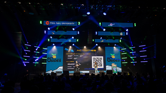 Two speakers stand on a stage in front of a large screen with the BSides Orlando logo displayed. Above it another screen displays the Full Sail University logo.