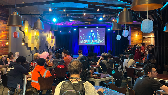 A crowd of students sit at tables in a large room. A large screen displays a video game and decorative paper lanterns hang around the room.