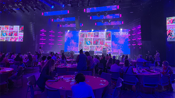 Groups of people sit around circular tables in a large room with pink and blue lights. A large screen displays photos and the American Cancer Society Making Strides Against Breast Cancer logo