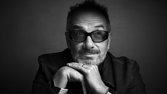 A black and white portrait of singer, songwriter, and producer Elvis Costello wearing dark-tinted glasses and a wedding band.