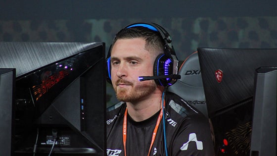 A focused esports player wearing a headset competes at a gaming event, seated between two monitors with a dark, intense atmosphere.