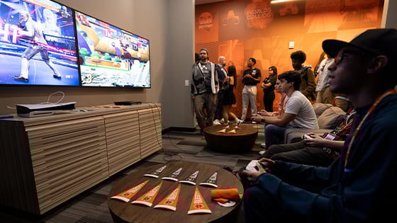Gamers compete in a lounge with dual screens, a PlayStation 5, and Armada-branded decor, while a crowd watches in the background.