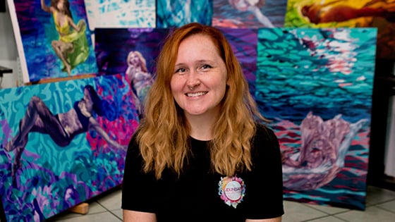 Jessica sits in front of several of her colorful paintings. She is wearing a black t-shirt and is smiling.