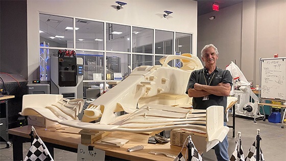 Course Director Pat Starace, a man with white hair and a grey polo shirt, standing next to a large workbench with a 3D-printed replica of a Formula 1 race car in Full Sail’s SimLab.