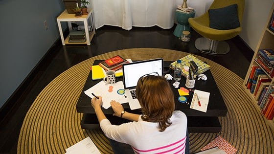 A student sits cross-legged on the floor in front of a coffee table with a laptop, papers, notebooks, and pens.