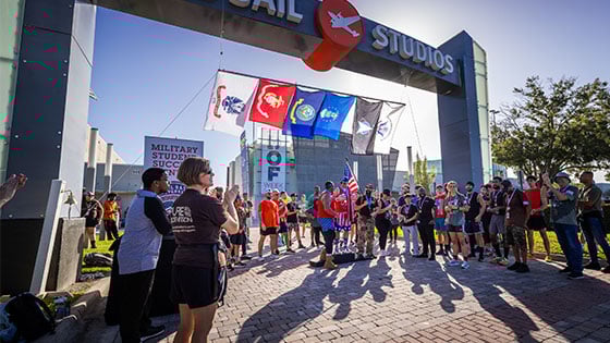 Three 5K winners stand on a podium with medals around their necks under the Full Sail Studios archway. Other competitors stand near them and applaud.