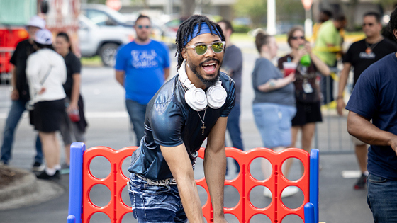 A student smiles while wearing headphones around their neck, sunglasses, a blue headband, and a dark shirt. A giant Connect 4 game and a crowd of people are behind them. The words “Student Community Association Throws Campus Life Block Party” are displayed on the photo.