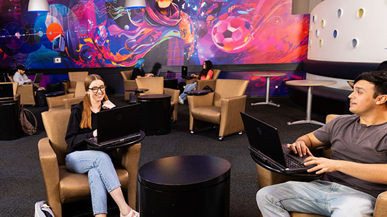 Students study and relax with laptops in Full Sail’s 3B Art Building Lobby, featuring colorful mural art and lounge seating.