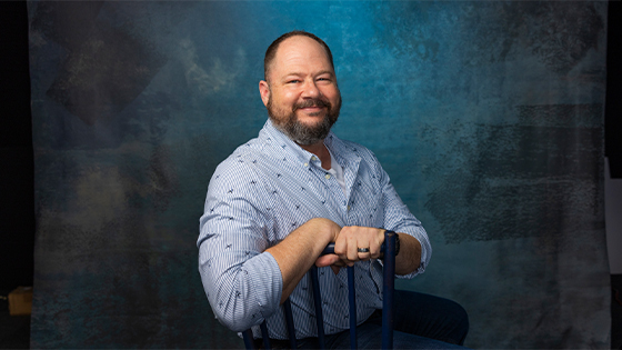 A man with glasses, short dark hair, and a beard is seen wearing a white shirt with a small animal pattern while smiling against a beige wall.
