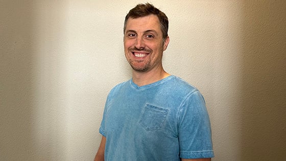 James Lane stands in front of a beige wall and smiles at the camera. He is wearing a light blue v-neck t-shirt.