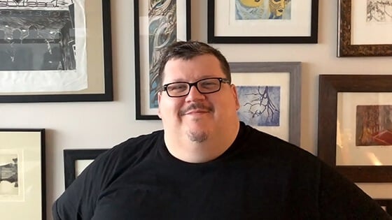 A smiling man wearing glasses and a black t-shirt sits in front of a wall covered with framed paintings.