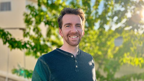 Lee Anderson stands outdoors in front of a tree and smiles at the camera. He has red hair and a beard, and he’s wearing a teal henley.