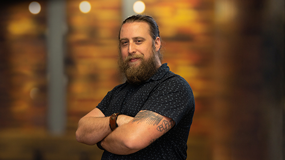 Kyle is standing with his arms crossed, smiling gently. He is wearing a short sleeve blue shirt and his hair is in a bun.