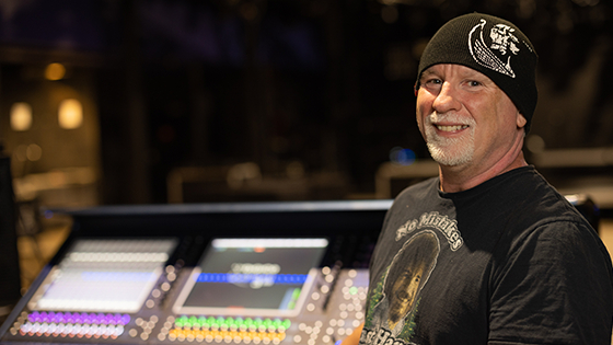 Gregg in a sound studio wearing a black beanie and graphic t-shirt. He is smiling.